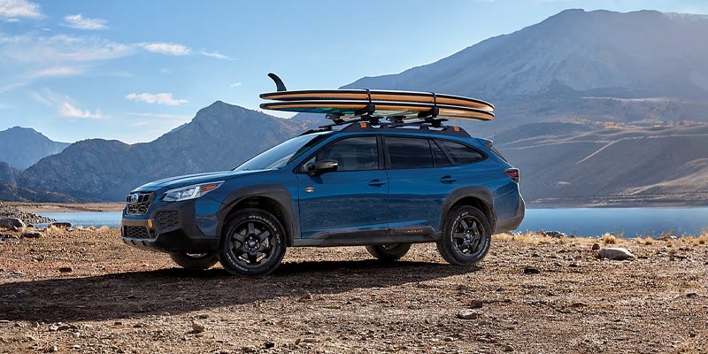 A blue Subaru Outback with roof racks holding surfboards, parked in a rugged outdoor setting with mountains and a lake in the background.