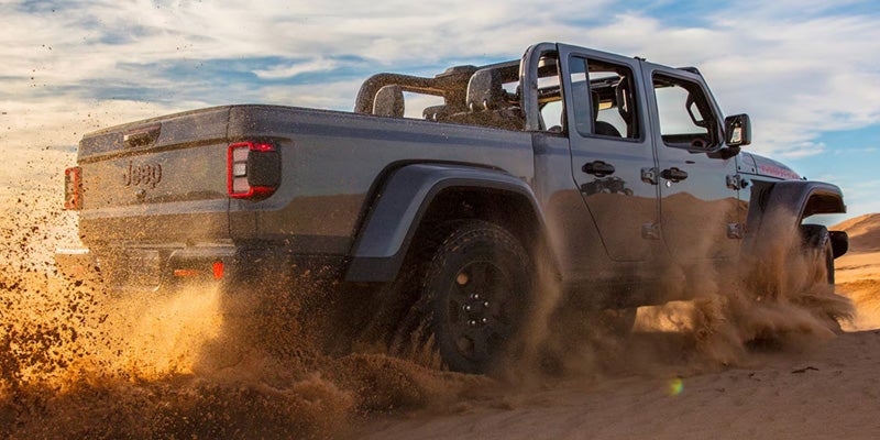 A 2026 Jeep Gladiator truck driving through sandy terrain, kicking up dust and sand.