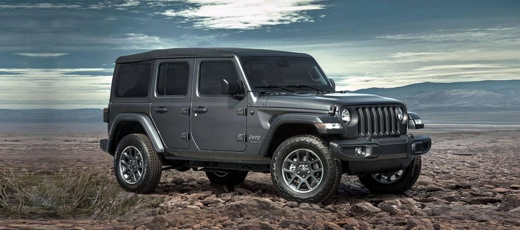 A gray Jeep Wrangler parked in a rugged, outdoor landscape with mountains in the background under a cloudy sky.