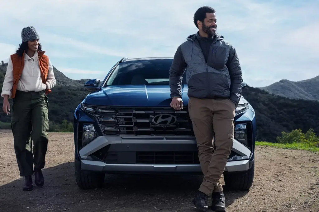 A man and a woman standing next to a blue Hyundai vehicle in a mountainous outdoor setting, with the woman smiling and the man leaning against the car.