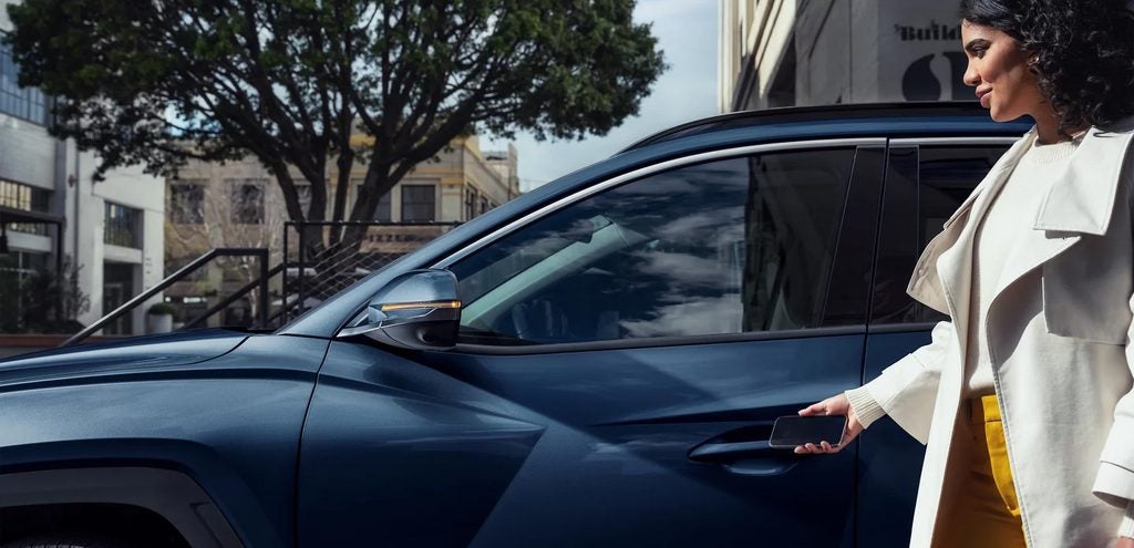 A woman holding a smartphone stands beside a blue SUV, preparing to open the driver’s door on a city street.
