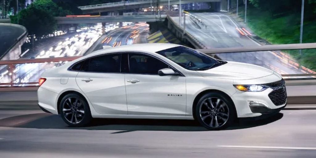 A white 2023 Chevrolet Malibu driving on a road with a fountain behind it.