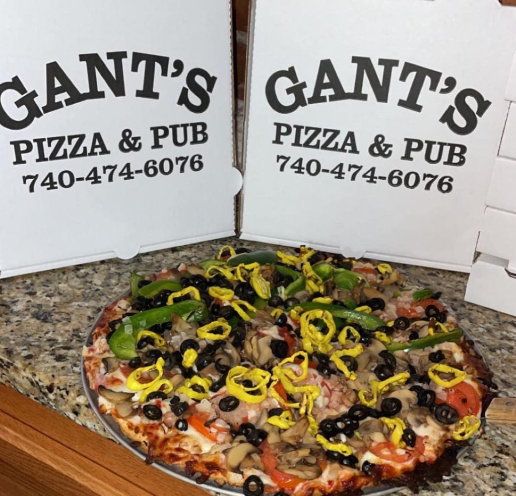 A photo of a loaded pizza with various toppings in front of two cardboard pizza boxes that say "Gant's Pizza &amp; Pub" with their phone number on it: 740-474-6076.