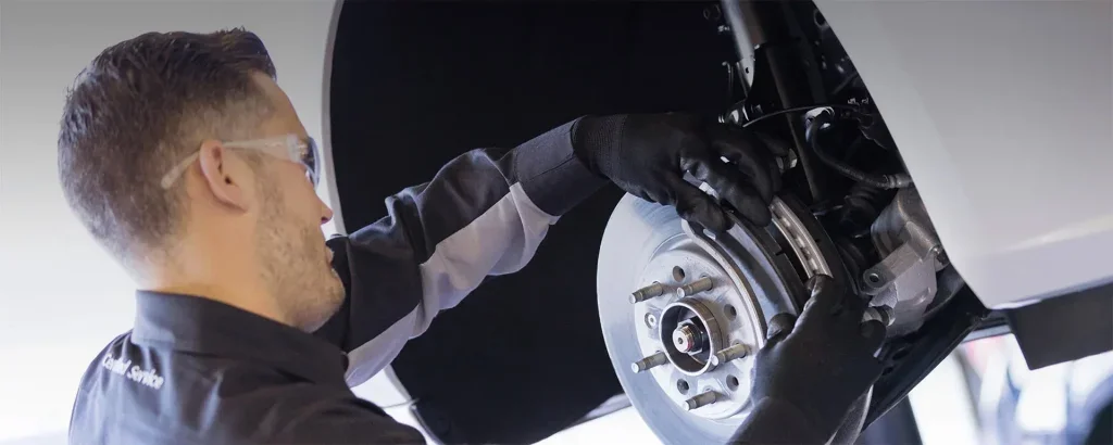 Technician performing brake service on a vehicle wheel hub with rotor and caliper exposed in an auto repair shop.
