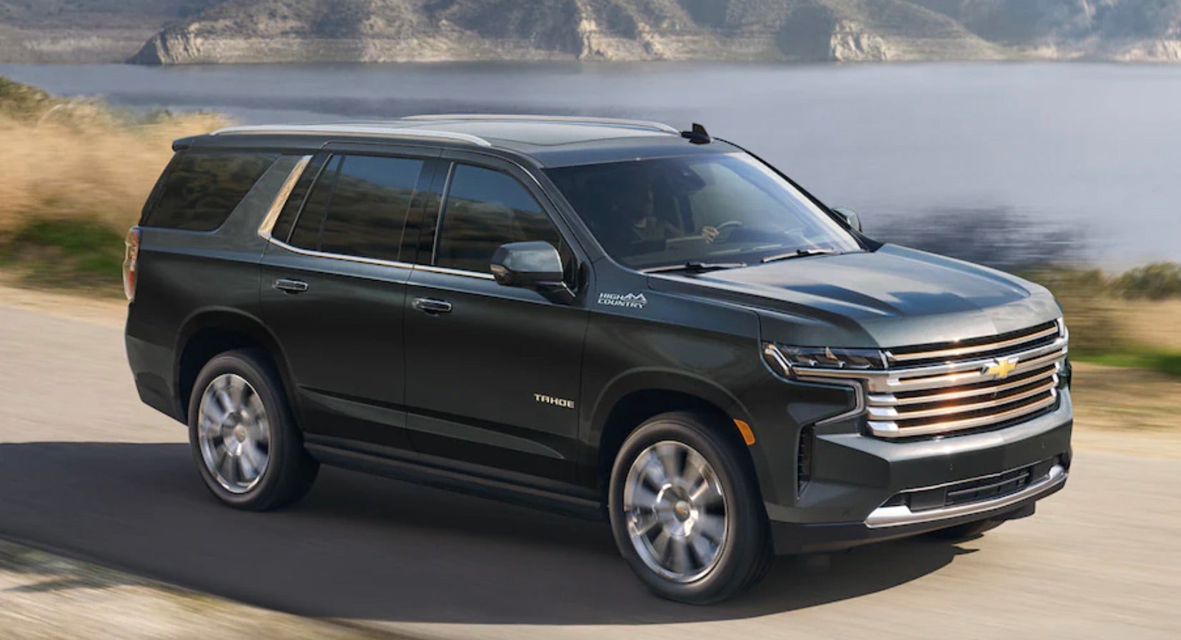 2022 Chevy Tahoe driving along the waters edge with the sun beaming down and the mountains in the background.
