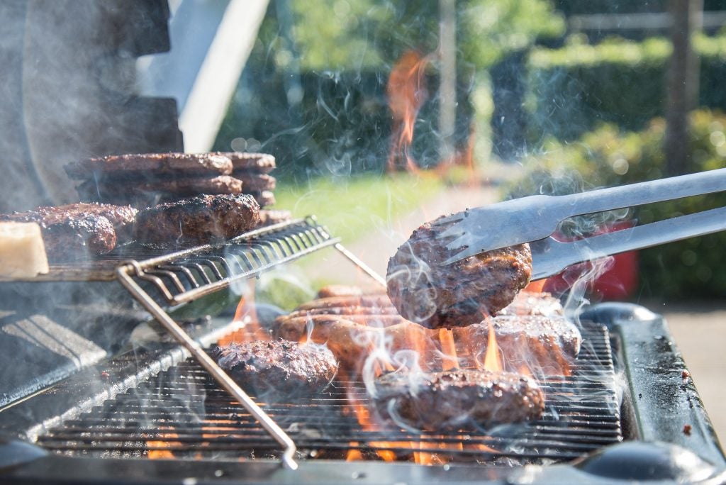 close up shot of someone grilling burgers flipping one over to cook the other side. There is a bunch of smoke around all burgers. 