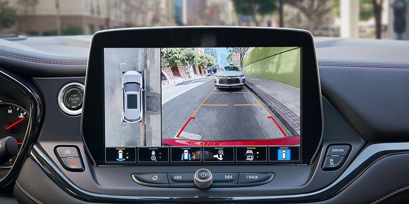 Interior view of a car dashboard showing a rear-view camera and 360-degree surround view display with parking guidelines and a street scene behind the vehicle for assisted parking.
