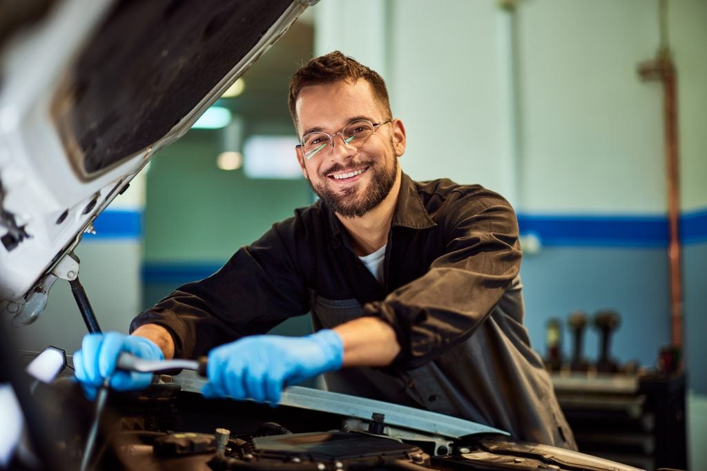 Smiling automotive mechanic wearing gloves and glasses working under the open hood of a car in a repair shop.