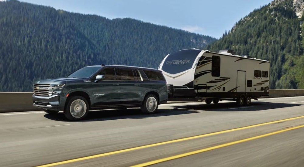 A blue 2023 Chevy Suburban High Country is shown towing a trailer on a mountain highway.