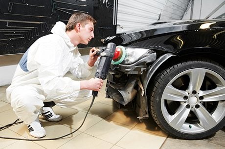 A technician wearing white protective overalls crouches down to buff the front bumper and headlight of a black car using a rotary polisher inside a body shop.