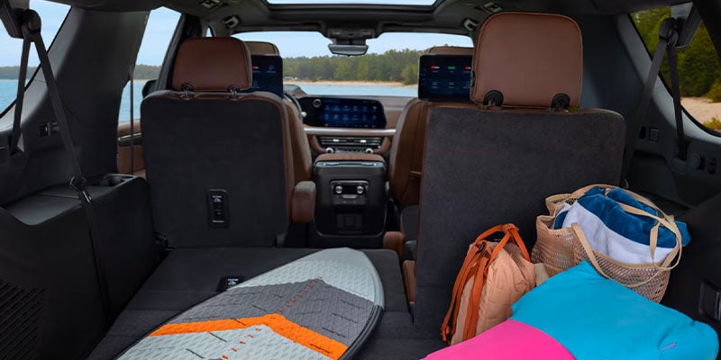 The back of an 2025 Chevy Suburban SUV showing a surfboard, colorful bags, and screens in front seats.