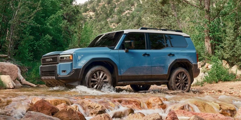 Blue Toyota SUV driving through a rocky stream in a forest landscape.