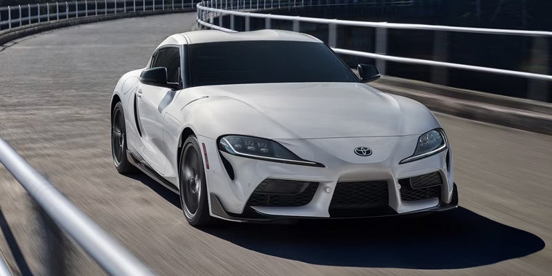 A white Toyota Supra speeding along a curved road with a railing on the side.