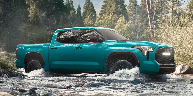 Teal Toyota pickup truck driving through a shallow stream in a forested area.