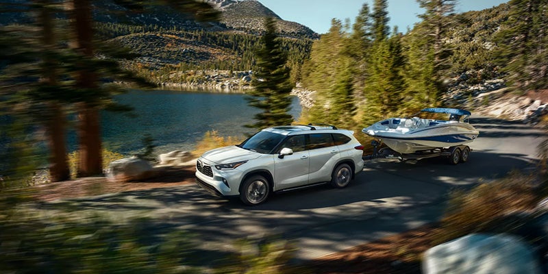 White Toyota SUV towing a boat along a scenic lakeside mountain road.