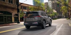 A gray Toyota RAV4 driving through a city street with tall buildings and green trees lining the road.