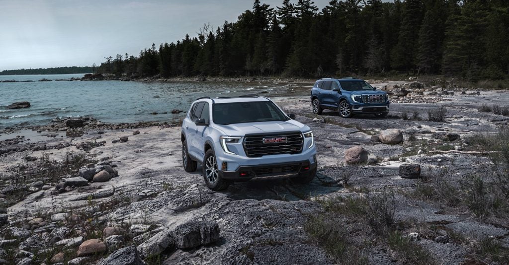 Two GMC SUVs parked on a rocky lakeshore with pine trees and overcast skies in the background.