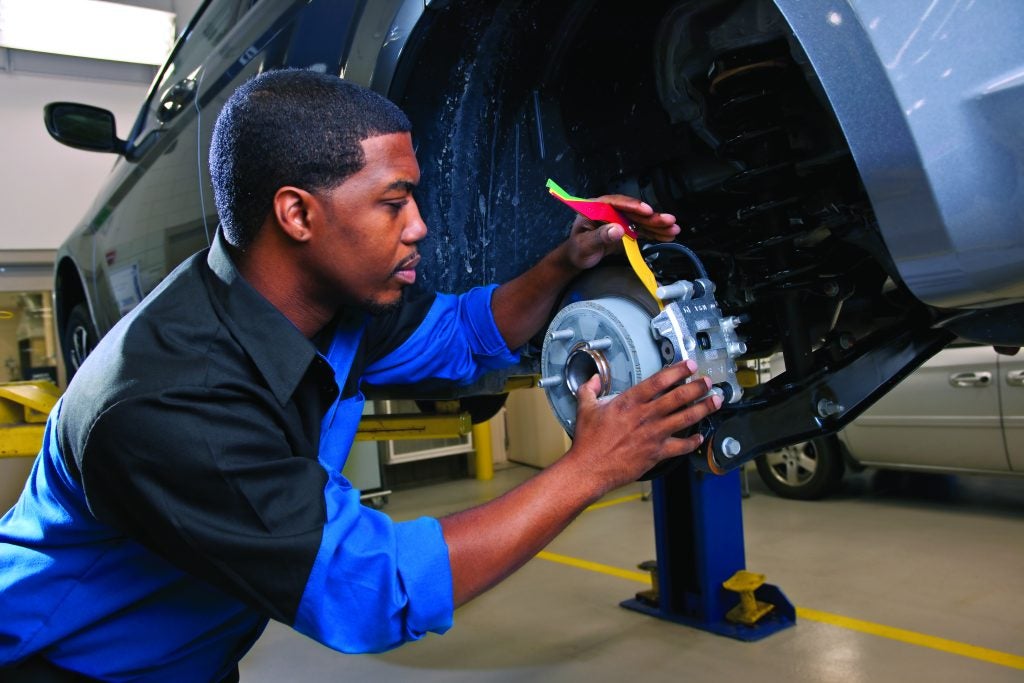 A mechanic in a blue and black uniform uses a color-coded brake lining thickness gauge to inspect the brake pads on a vehicle lifted on a shop hoist.