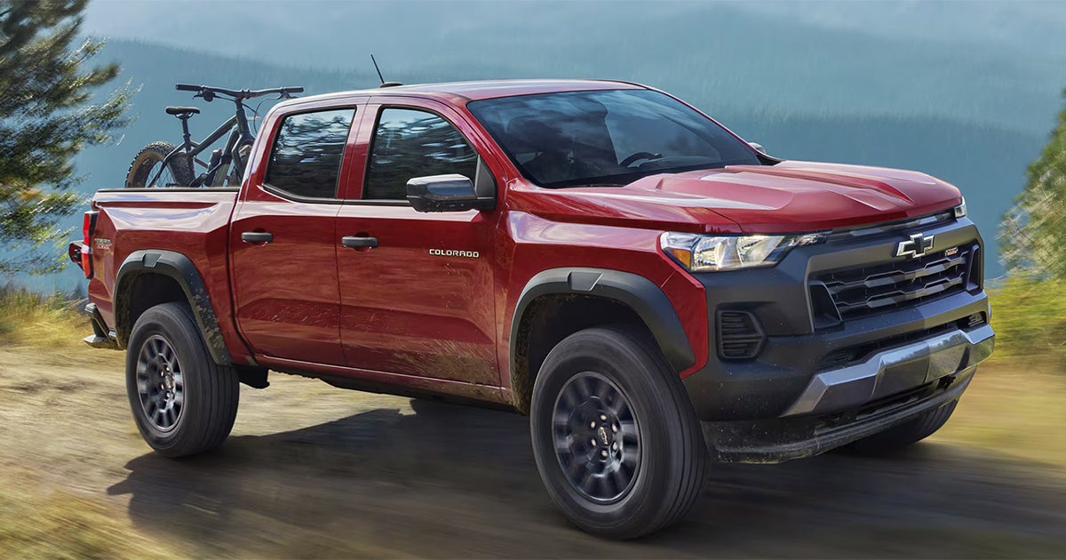 Red Chevrolet Colorado pickup truck with mountain bikes in bed parked near lake with mountains in background