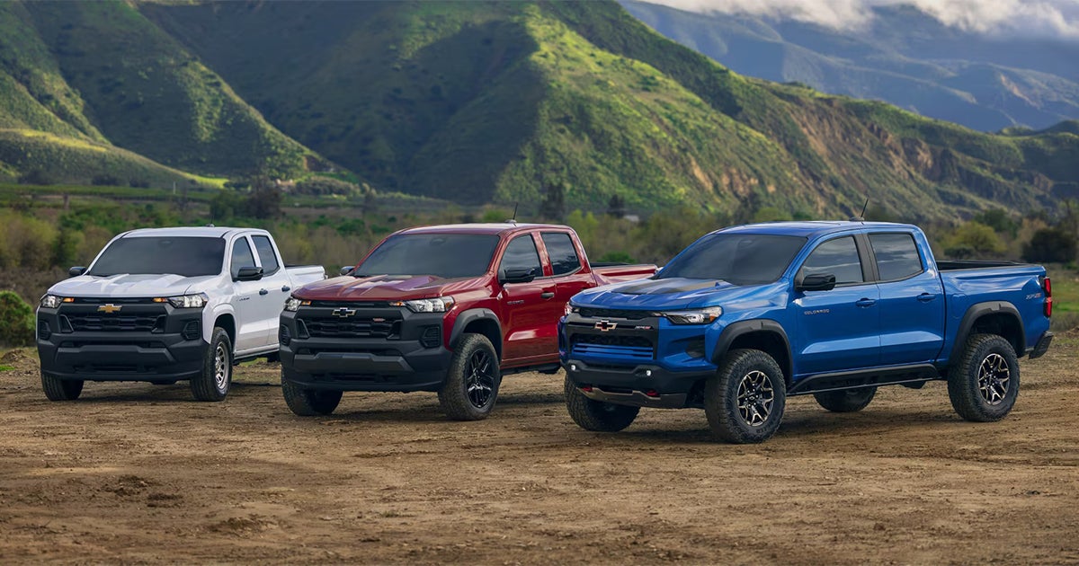 Three Chevrolet Colorado pickup trucks in white, red, and blue parked side by side with mountain backdrop