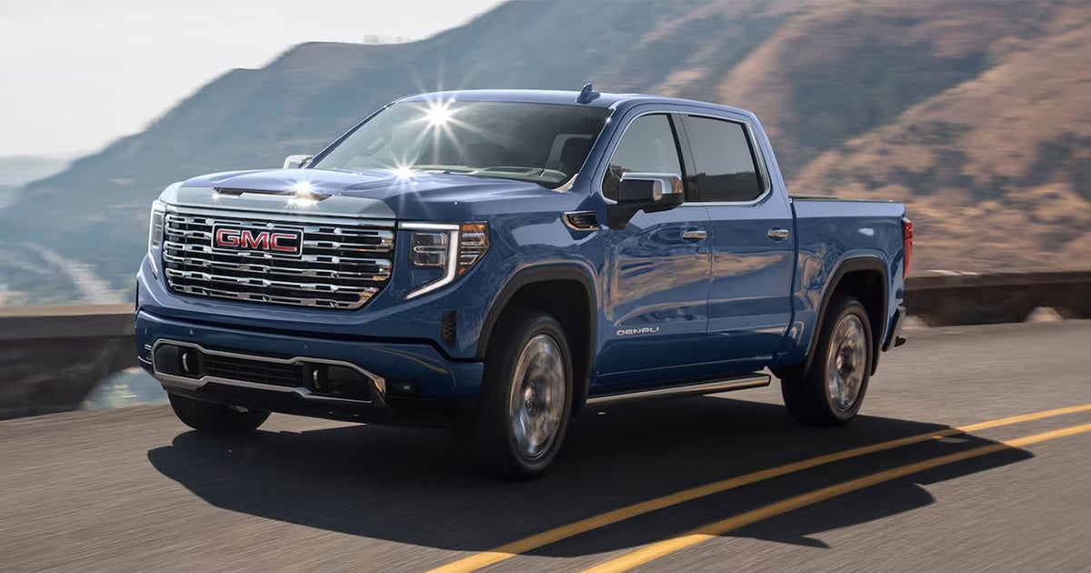 Blue GMC Sierra Denali driving along a winding mountain road, with sunlight reflecting off its chrome grille and accents.