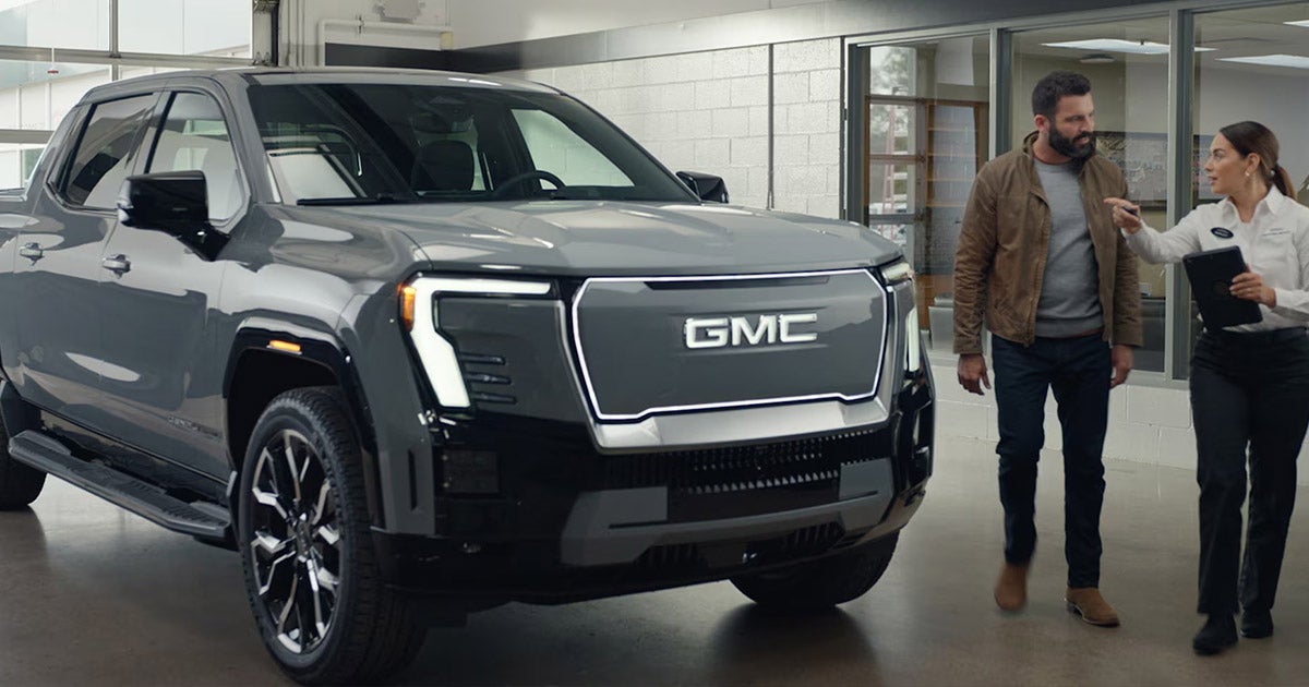 Customer and dealership representative standing beside a silver GMC Sierra EV inside a showroom, with the representative holding a tablet and pointing while speaking to the customer, highlighting the truck’s illuminated GMC grille and modern styling.