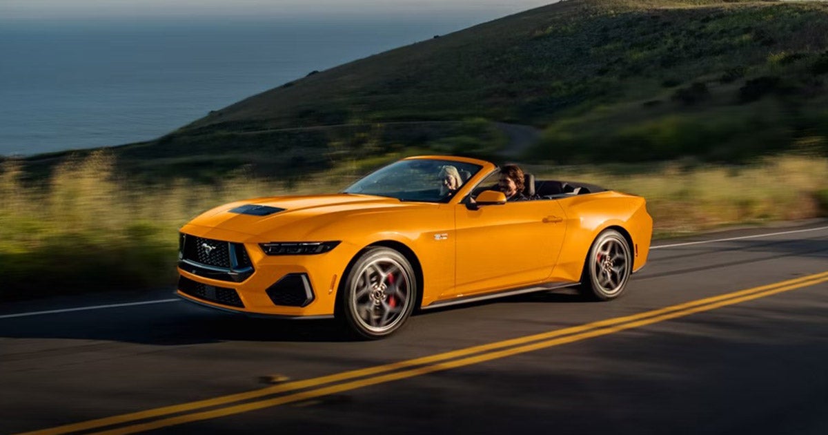 2025 Ford Mustang GT convertible in bright orange driving along a scenic coastal highway, featured in South Bay Ford’s review comparing the GT, EcoBoost, and Dark Horse models near Los Angeles, CA.