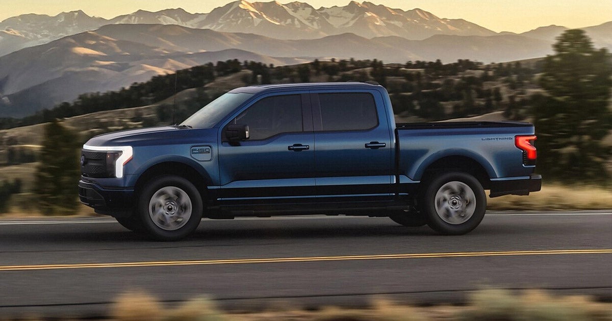 A blue Ford F-150 Lightning pickup truck travels on a road with a mountainous landscape in the background. The scene conveys a sense of adventure and freedom.