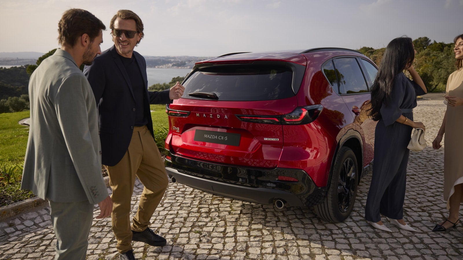 A group of people stand cheerfully around a shiny red Mazda CX-5 on a cobblestone path, overlooking a scenic landscape with distant hills and water.