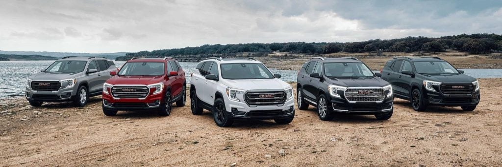 Lineup of the 2026 GMC SUV models sitting on the lakeside shore on a blue-grey sky day with low mountains in the background