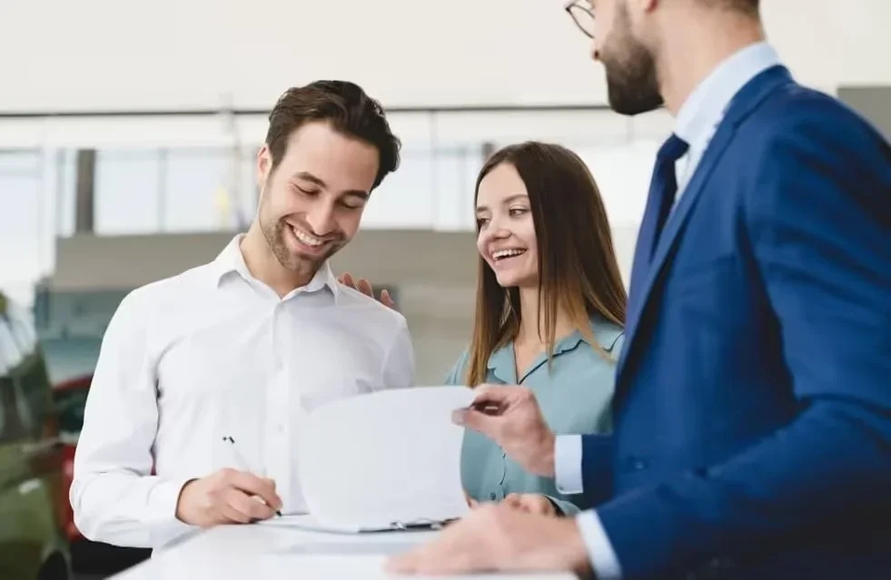 A smiling couple sign paperwork for buying a new GMC SUV with smiling salesman looking on