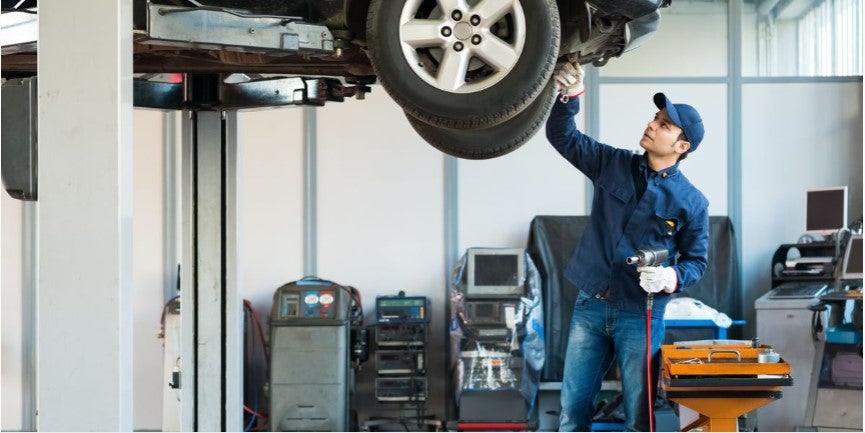 Mechanic working in an automotive repair shop inspecting or servicing a vehicle lifted on a hydraulic lift, while holding tools and examining the wheel area.