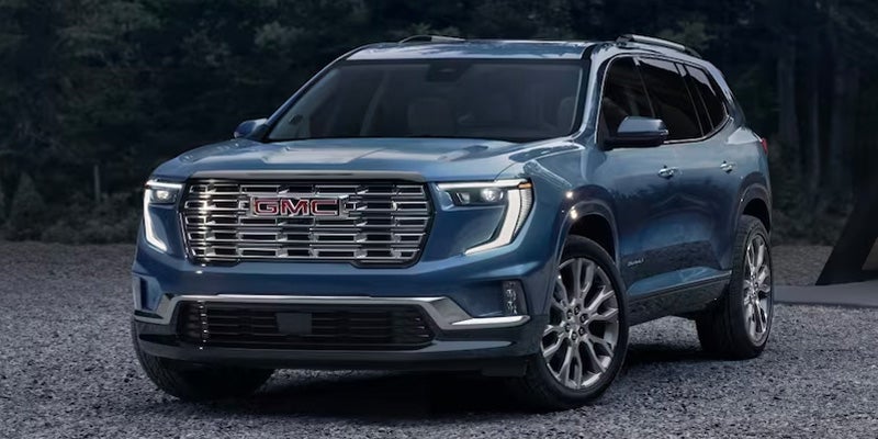 Front three-quarter view of a used blue GMC SUV parked outdoors on a gravel surface, featuring a bold chrome grille, LED headlights, and alloy wheels.