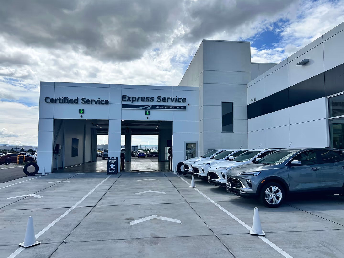 Car dealership service center with Express Service and Certified Service bays, white vehicles parked, cloudy sky backdrop