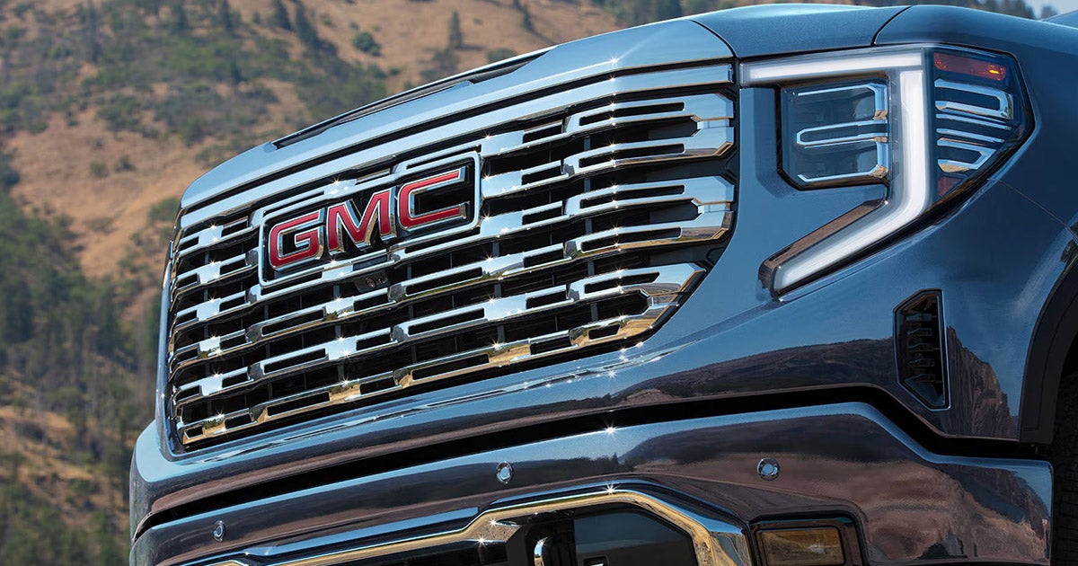 GMC Sierra 2024 chrome grille and headlight, blue truck with mountainous terrain background