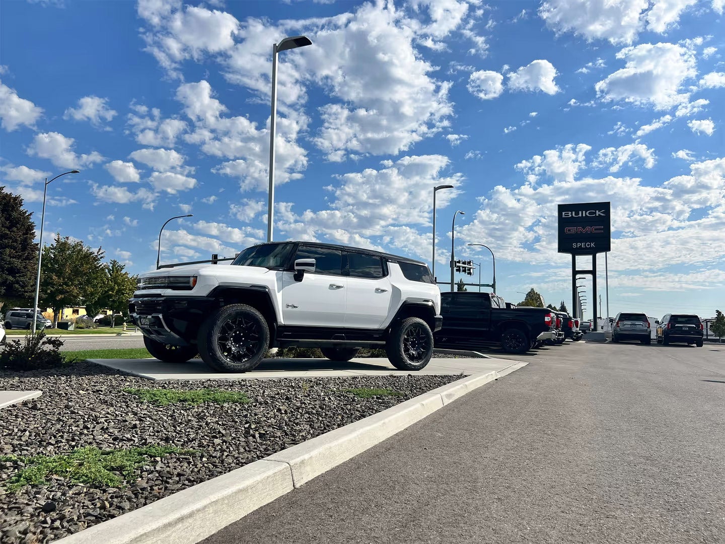 White GMC Hummer EV SUV parked at Buick dealership on cloudy day with blue sky and street lamps