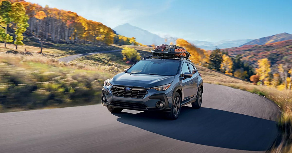 Subaru Crosstrek driving on winding mountain road with autumn trees, roof cargo box, scenic landscape background