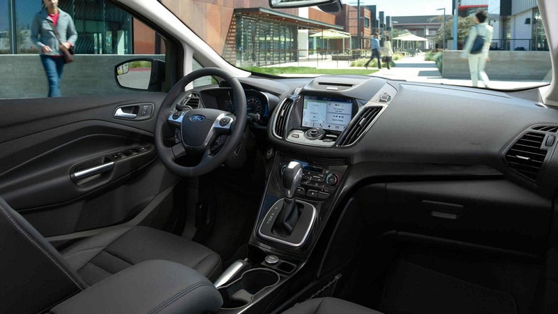 Driver's seat view of a black car interior, dashboard, and touchscreen, with people walking outside.