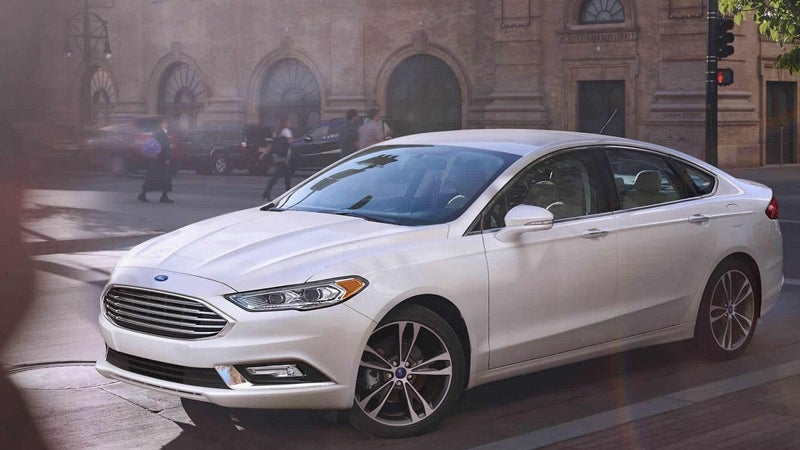 A white Ford Fusion sedan parked on a city street with historical buildings and blurred people in the background.