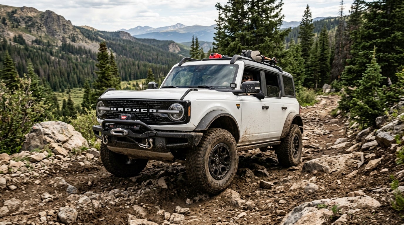 A white Ford Bronco drives over a rocky, dirt off-road trail in a pine forest mountain setting. (Representative image for illustration purposes only. Actual vehicle may vary based on trim and configuration.)