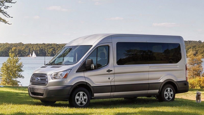 A silver Ford Transit passenger van parked on a grassy patch near a lake with sailboats and a forested shore.