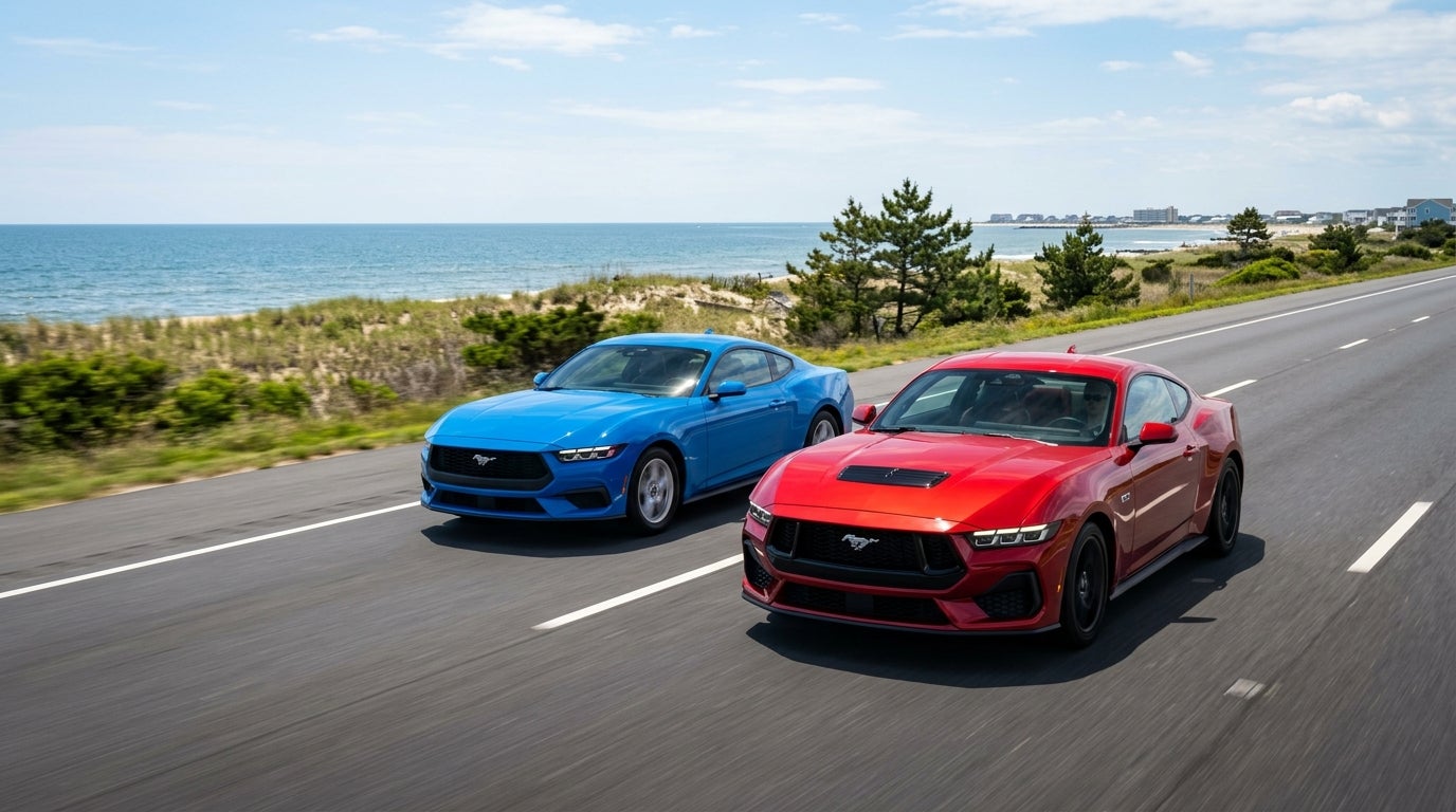 A red Ford Mustang and a blue Ford Mustang drive side-by-side on a scenic coastal highway next to the ocean. (Representative image for illustration purposes only. Actual vehicle may vary based on trim and configuration.)