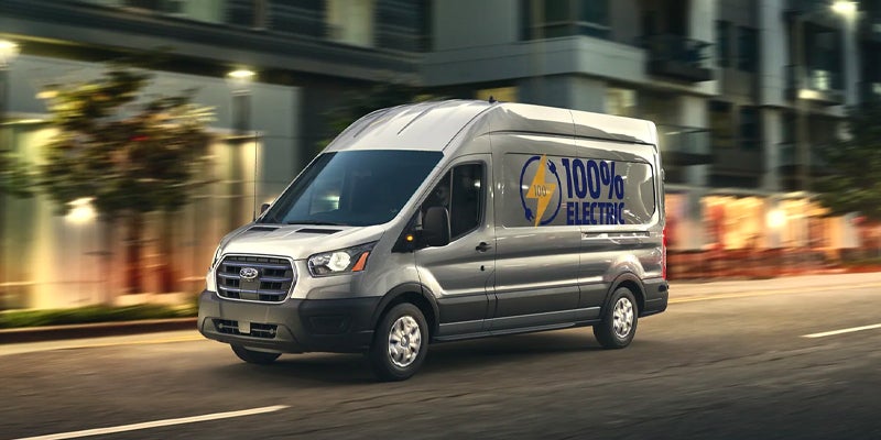 A gray Ford Transit cargo van with a "100% Electric" lightning bolt decal speeds down a city street.