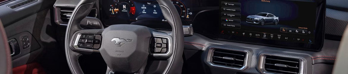 Interior of a Ford Mustang, showing the steering wheel with Mustang logo, digital instrument cluster, and large infotainment screen displaying a gray Mustang.