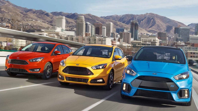 Three Ford Focus models in red, yellow, and blue drive together on a highway with a city and mountains beyond.