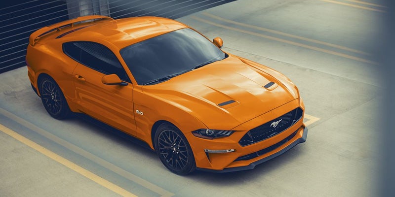 An orange Ford Mustang sports car with black wheels and spoiler, parked in a concrete lot.