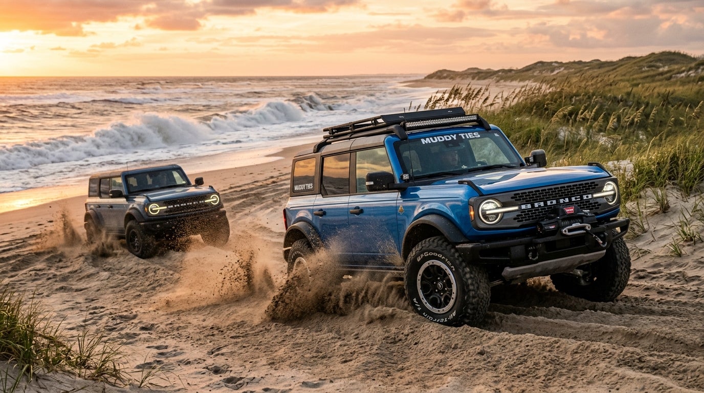 Blue Ford Bronco driving through sandy beach dunes near ocean waves at sunset with another Bronco following. (Representative image for illustration purposes only. Actual vehicle may vary based on trim and configuration.)