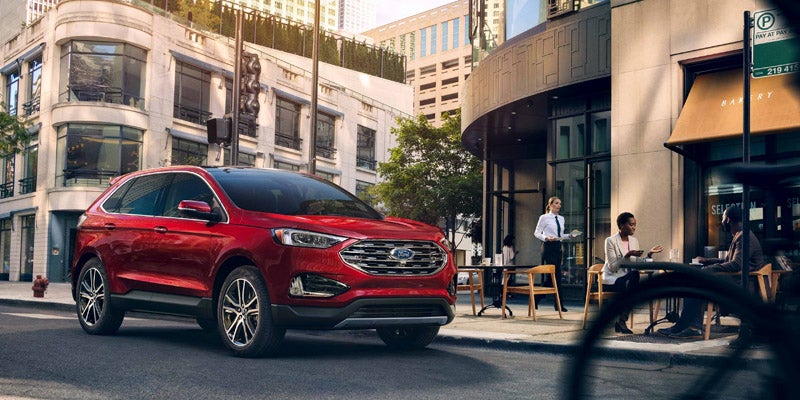 A red Ford SUV is parked on a city street next to a cafe with people sitting outside.