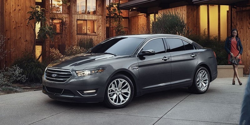 A dark gray sedan parked on a concrete driveway in front of a modern wooden house, with a woman standing nearby.