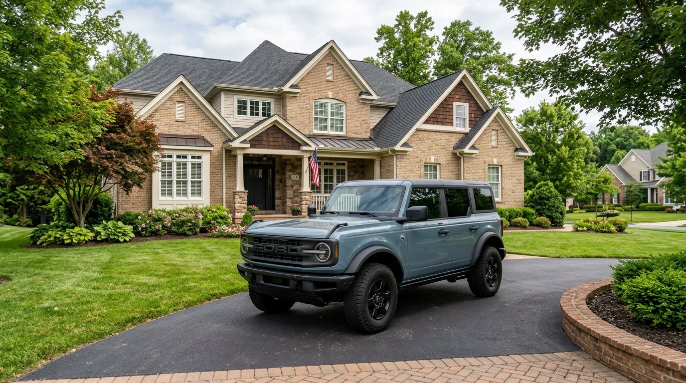 Blue-gray Ford Bronco SUV parked on a paved driveway in front of a large two-story brick suburban home. (Representative image for illustration purposes only. Actual vehicle may vary based on trim and configuration.)